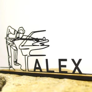 May include: A black metal sculpture of a person playing pool, with the name "ALEX" in block letters. The sculpture is mounted on a wooden base and sits on a stone surface. The design is a minimalist, line-art style.