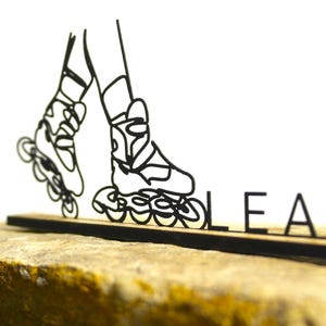Peut inclure: Dessin au trait noir de patins à roulettes avec le nom "LEA" découpé en lettres noires. Les patins sont sur une base en bois, sur fond blanc. La base repose sur une surface texturée et beige.