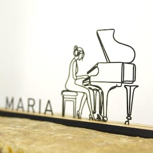 Pode incluir: Uma placa de nome de mesa de madeira cortada a laser com o nome "MARIA". Um desenho a traço preto de uma pessoa tocando um piano de cauda está à direita. A placa de nome está em uma superfície de pedra.