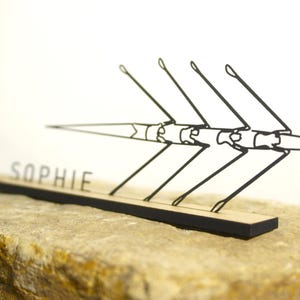 Peut inclure: Sculpture en métal noir représentant un bateau d'aviron avec quatre rames, montée sur une base en bois. Le nom "SOPHIE" est découpé au laser dans la base. La sculpture est posée sur un fond blanc et repose sur une surface en pierre.