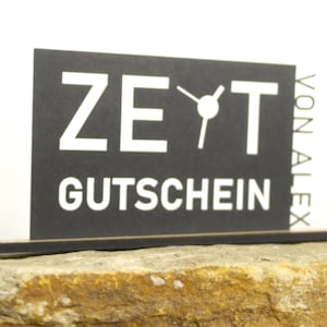 Könnte beinhalten: Eine schwarze rechteckige Karte mit dem Wort "ZEIT" über "GUTSCHEIN" in Weiß. Die Karte hat eine Grafik einer Uhr und die Worte "VON ALEX" auf der rechten Seite. Die Karte liegt auf einem strukturierten, hellbraunen Felsen.