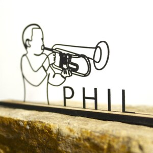 Op de afbeelding: Een zwarte lijntekening van een persoon die een trombone bespeelt. De tekening is gemonteerd op een houten voet met de naam "PHIL" in afzonderlijke letters. De voet rust op een getextureerd, bruinachtig oppervlak.