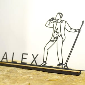 May include: A black laser-cut metal nameplate with the name "ALEX" and a singer figure. The singer is holding a microphone and standing on a stage. The nameplate is on a wooden base.