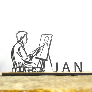 Könnte beinhalten: Eine schwarze, lasergeschnittene Holzskulptur eines Künstlers, der auf einer Leinwand malt, mit dem Wort "JAN" daneben. Der Künstler sitzt und hält einen Pinsel. Die Skulptur steht auf einer Steinoberfläche.