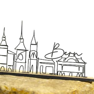 Könnte beinhalten: Eine schwarze Drahtskulptur der Skyline von Bern, die Wahrzeichen und den Namen der Stadt zeigt. Das Kunstwerk ist auf einem Holzsockel montiert und auf einer Steinoberfläche ausgestellt.