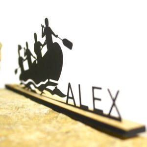 Puede incluir: Silueta negra de una escena de rafting con el nombre "ALEX" recortado en madera. El diseño presenta una balsa con personas remando, colocada sobre una base de madera. El fondo es blanco y la base está sobre una superficie marrón.