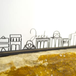 Puede incluir: Silueta de metal negro del horizonte de Jericó, con varios contornos arquitectónicos. La palabra "Jericho" está escrita en cursiva sobre el horizonte. La obra de arte se muestra sobre una roca marrón claro sobre un fondo blanco.