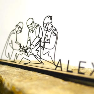 Puede incluir: Una escultura de alambre negro que representa tres figuras, posiblemente soldados, sobre una base de madera. El nombre "ALEX" está escrito en letras individuales. La escultura está sobre una superficie texturizada de color marrón claro.
