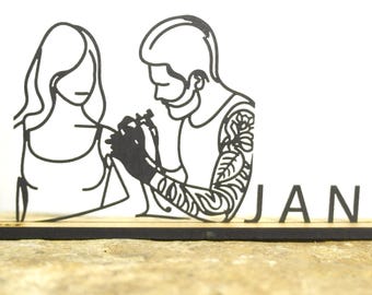 May include: A black line art illustration of a tattoo artist giving a tattoo to a person. The word "JAN" is displayed in black letters on a wooden base. The figures are on a light background.