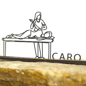 Puede incluir: Ilustración en blanco y negro de un masajista tratando a un paciente en una camilla. La palabra "CARO" se muestra en letras negras sobre una base marrón. La imagen está sobre un fondo blanco y descansa sobre una superficie de piedra.