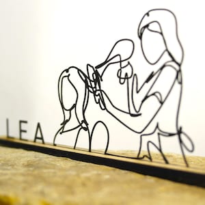 Puede incluir: Escultura de alambre negro que representa a un peluquero con un cliente, sosteniendo un secador de pelo y unas tijeras. La palabra "LEA" está escrita en letras negras individuales sobre una base de madera. La escultura está sobre un fondo neutro.