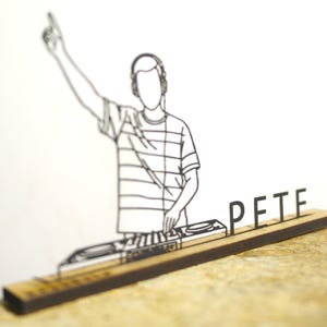 Könnte beinhalten: Schwarz-weiße Strichzeichnung eines DJs mit Kopfhörern und einem gestreiften Hemd, der einen Arm hebt. Der Name "PETE" wird in schwarzen Buchstaben auf einem Holzsockel mit einem Plattenspieler angezeigt.