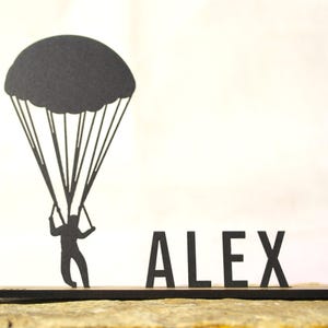 Op de afbeelding: Een zwarte silhouet-uitsparing met een persoon die met een parachute afdaalt, bevestigd aan een horizontale basis, met de naam ALEX in dikke hoofdletters naast de figuur, alles vervaardigd uit een plat materiaal.