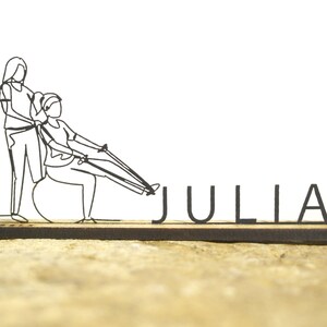 Può includere: Una scultura in filo nero raffigurante due figure che si esercitano, con il nome "JULIA" in lettere nere. Le figure sono impegnate in una routine di allenamento, con una seduta su una palla e che utilizza fasce di resistenza. La scultura è montata su una base di legno.