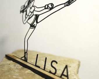 Könnte beinhalten: Eine schwarze Drahtskulptur einer Frau in einer Kampfsportpose, die tritt. Die Skulptur ist auf einem Holzsockel mit dem ausgeschnittenen Namen "LISA" montiert. Die Skulptur steht auf einem hellbraunen Stein.