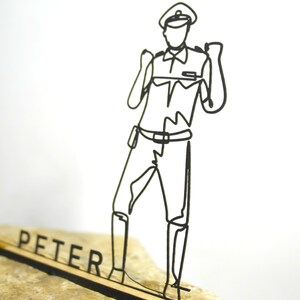 Könnte beinhalten: Eine schwarze Drahtskulptur eines Polizisten in Uniform, der auf einem Holzsockel mit dem Namen "PETER" in schwarzen Buchstaben steht. Die Skulptur steht vor einem weißen Hintergrund und einem beigen Stein.