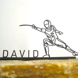 Op de afbeelding: Zwarte metalen sculptuur van een schermer in actie, met de naam "DAVID" aan de basis. Het kunstwerk toont een gestileerde omtrek van een schermer in een dynamische pose, die een degen vasthoudt. De sculptuur staat tegen een neutrale achtergrond.