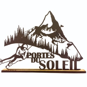 Op de afbeelding: Een zwart metalen silhouet van een skiër die een berg afdaalt met de woorden "Portes du Soleil" eronder. Het silhouet is gemonteerd op een houten basis.