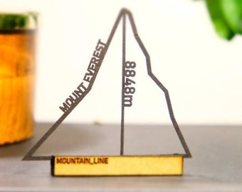 May include: A black and white paper model of Mount Everest with the text "Mount Everest" and "8848m" on the side. The model is sitting on a wooden surface with the text "Mountain Line" on a small wooden plaque.
