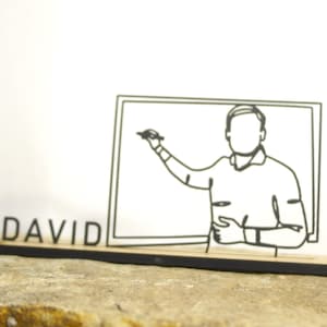 Könnte beinhalten: Ein Schreibtisch-Namensschild aus Holz mit dem Namen "DAVID" in schwarzen Buchstaben. Das Namensschild zeigt eine Strichzeichnung einer Person, die auf einer Tafel schreibt. Das Design ist minimalistisch und modern.