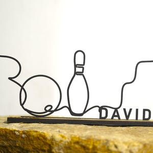 Könnte beinhalten: Schwarze Metallskulptur zum Thema Bowling mit Bowlingkugel, Kegel und dem Namen "DAVID". Das Design ist eine durchgehende Linie, montiert auf einem rechteckigen Sockel. Die Skulptur wird auf einer strukturierten, hellbraunen Oberfläche präsentiert.