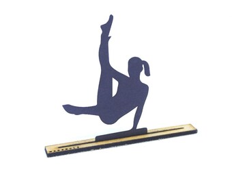 Könnte beinhalten: Eine schwarze Silhouette einer Turnerin, die einen Handstand auf einer Holzbasis macht. Die Basis ist mit "MADE SOLELY" beschriftet.