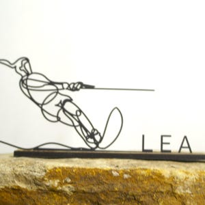 May include: Black metal sculpture of a waterskier in motion. The sculpture is mounted on a rectangular base, with the name "LEA" in block letters. The sculpture is placed on a rock.