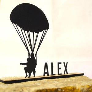 Könnte beinhalten: Eine schwarze Tischdekoration in Silhouettenform, die eine Person an einem Fallschirm zeigt, mit dem Namen ALEX in Großbuchstaben neben der Figur. Das gesamte Design ist auf einer dünnen, flachen, rechteckigen schwarzen Basis montiert.