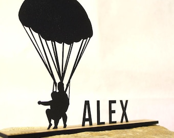Könnte beinhalten: Eine schwarze Tischdekoration in Silhouettenform, die eine Person an einem Fallschirm zeigt, mit dem Namen ALEX in Großbuchstaben neben der Figur. Das gesamte Design ist auf einer dünnen, flachen, rechteckigen schwarzen Basis montiert.