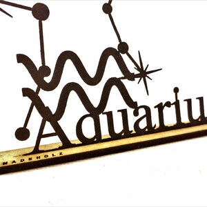 May include: Black wooden cutout of the Aquarius zodiac sign with a star and the word "Aquarius" written below. The cutout is on a wooden base with the words "MADEHOLZ" printed on the base.