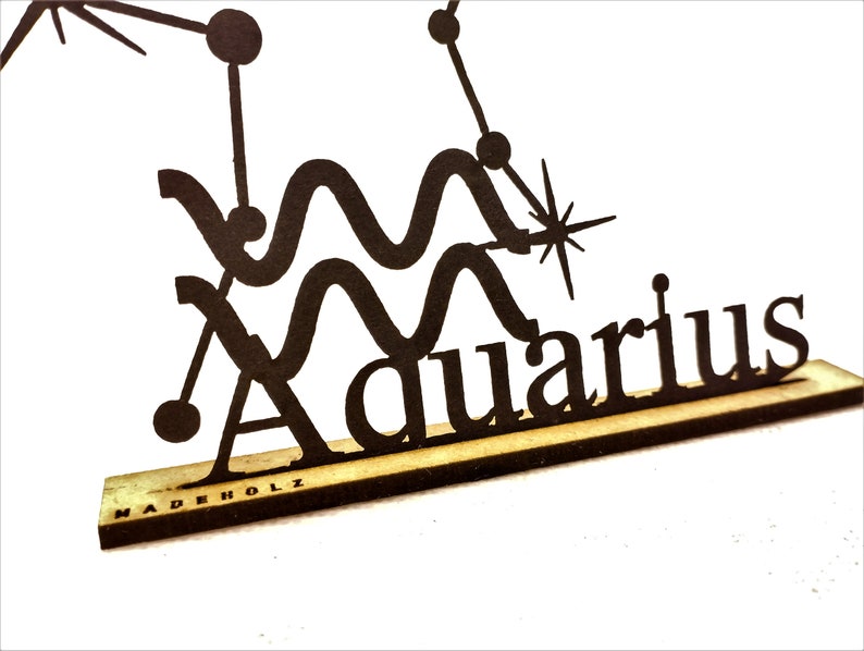 May include: Black wooden cutout of the Aquarius zodiac sign with a star and the word "Aquarius" written below. The cutout is on a wooden base with the words "MADEHOLZ" printed on the base.