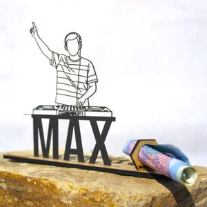 Op de afbeelding: Een zwart metalen silhouet van een DJ met een koptelefoon die achter een mengpaneel staat, met de naam MAX in grote hoofdletters eronder. Een kleine zeshoekige houten houder is aan de zijkant bevestigd en bevat een opgerold bankbiljet.