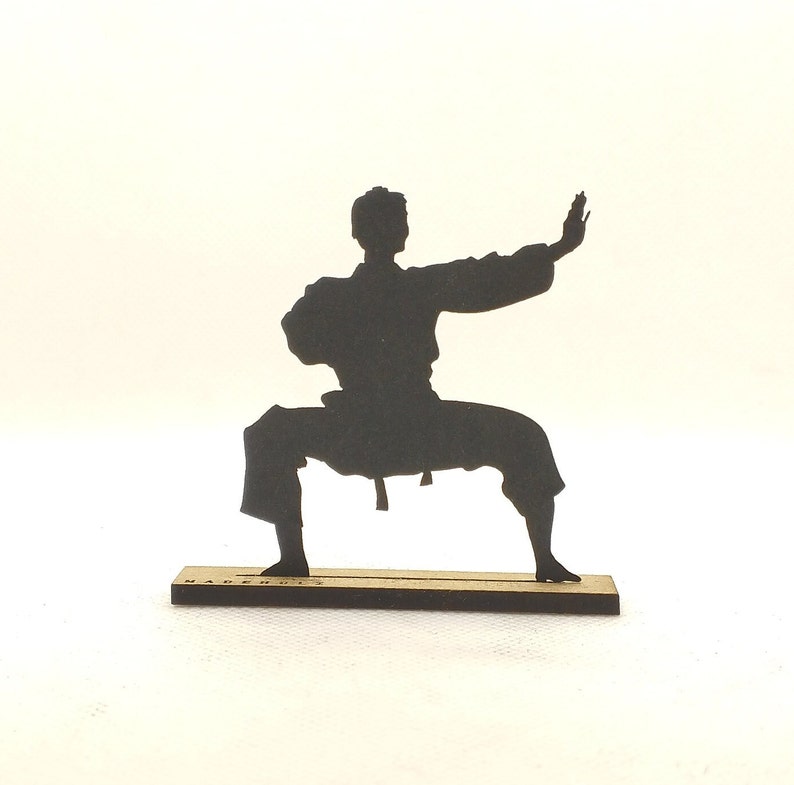 Pu&ograve; includere: Silhouette nera di una persona in posa di karate, in piedi su una base di legno. La base ha la scritta "MADE IN" stampata.