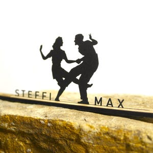 Könnte beinhalten: Eine schwarze Silhouette eines tanzenden Paares auf einem Holzsockel mit den ausgeschnittenen Namen "STEFFI" und "MAX". Der Sockel ruht auf einem strukturierten, beigefarbenen Stein vor weißem Hintergrund.