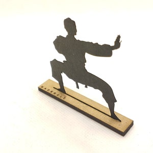 Pu&ograve; includere: Silhouette nera di una persona in una posa di arti marziali, in piedi su una base di legno con il testo "MADEHOLZ" inciso su di essa.