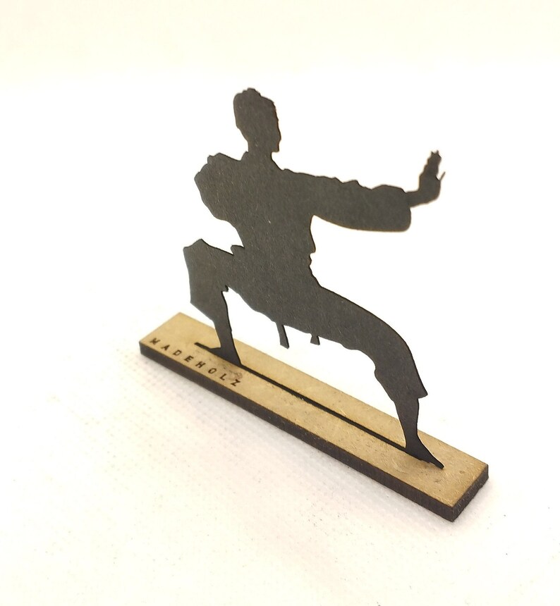 Pu&ograve; includere: Silhouette nera di una persona in una posa di arti marziali, in piedi su una base di legno con il testo "MADEHOLZ" inciso su di essa.