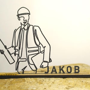 Puede incluir: Una figura de arte lineal negro de un trabajador de la construcción sosteniendo un plano, usando un casco y chaleco. El nombre "JAKOB" se muestra en una base de madera. El diseño es minimalista y moderno, adecuado para la decoración de escritorio.