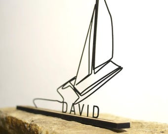 Puede incluir: Escultura de velero de metal negro con el nombre "DAVID" en letras negras sobre una base de madera. La escultura está sobre un fondo blanco y descansa sobre una superficie de piedra texturizada.