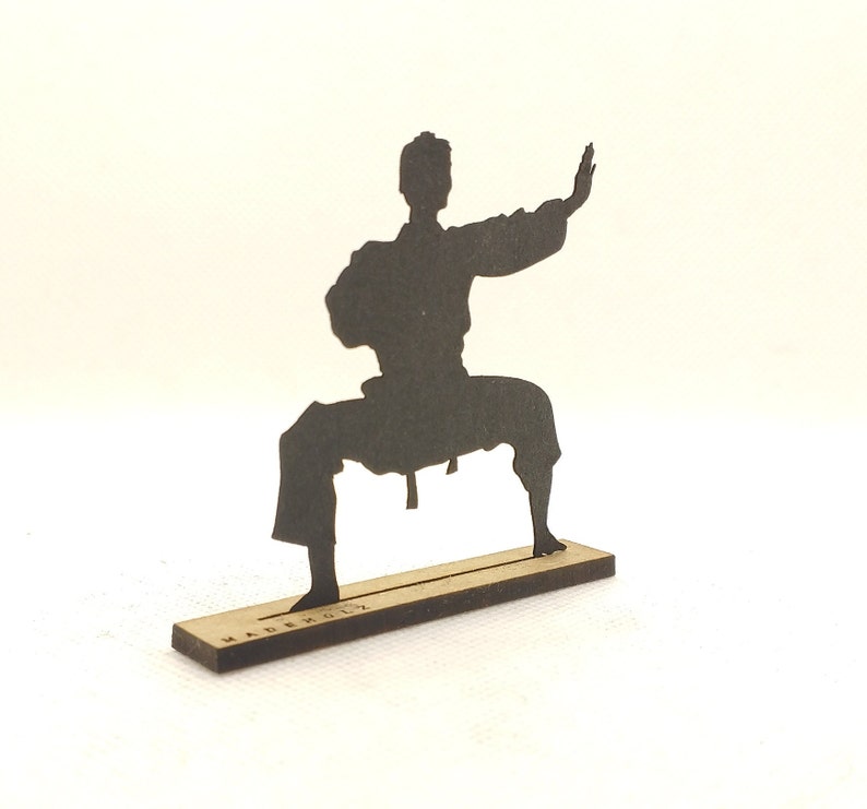 Pu&ograve; includere: Una silhouette nera di una persona in una posa di arti marziali, in piedi su una base di legno con la scritta "MADE IN ITALY" stampata su di essa.