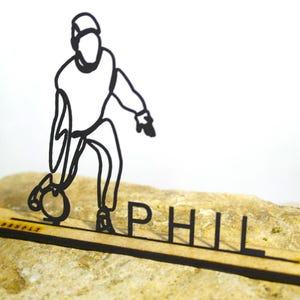 Könnte beinhalten: Eine schwarze Strichzeichnung eines Bowlingspielers in Aktion, der eine Bowlingkugel hält. Die Figur ist auf einem Holzsockel mit dem ausgeschnittenen Namen "PHIL" montiert. Der Hintergrund ist eine helle Oberfläche.