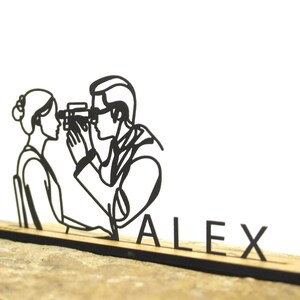 May include: A black laser-cut wooden desk accessory featuring a stylized optometrist examining a patient's eyes. The name "ALEX" is displayed in black letters on the base. The accessory is set against a neutral background.