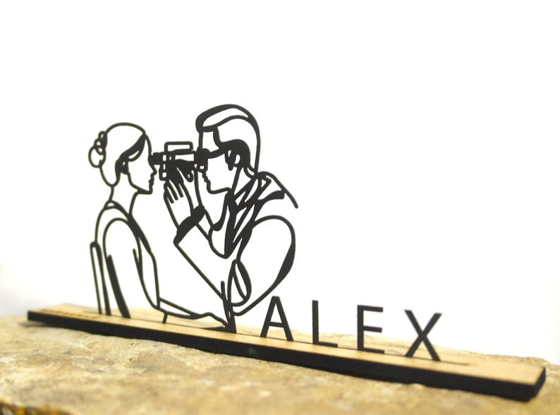 May include: A black laser-cut wooden desk accessory featuring a stylized optometrist examining a patient's eyes. The name "ALEX" is displayed in black letters on the base. The accessory is set against a neutral background.