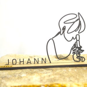 Op de afbeelding: Een lasergesneden houten bureau-accessoire met een minimalistische lijntekening van een persoon die een microscoop gebruikt. De naam "JOHANN" is eronder uitgesneden. Het accessoire wordt gepresenteerd op een lichtgekleurde steen.