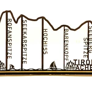 Könnte beinhalten: Eine schwarze Holzsilhouette einer Bergkette mit den Namen der Gipfel: Rofan Spitze, Seekarspitze, Hochiss, Barenkopf, Seeberg Spitze. Der Text "Tirol Achensee" befindet sich am Fuße der Berge.