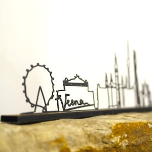 May include: A black metal skyline silhouette of a city, including a Ferris wheel and buildings, with the word "Viena" in cursive. The artwork is mounted on a rectangular base and sits on a stone surface.