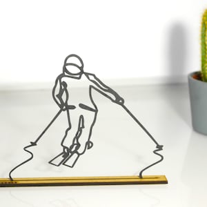 Peut inclure: Figurine de skieur en métal noir sur une base en bois. Le skieur est en mouvement, avec des bâtons et des skis. Un cactus dans un pot gris est en arrière-plan. Objet décoratif.