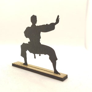 Pu&ograve; includere: Silhouette nera di una persona in una posa di arti marziali, in piedi su una base di legno.