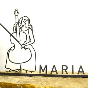Puede incluir: Una placa con el nombre de madera cortada con láser negro que presenta a un músico estilizado tocando el violonchelo. El nombre "MARIA" se muestra en letras negras individuales. La placa con el nombre está colocada sobre una superficie de piedra de color claro sobre un fondo blanco.