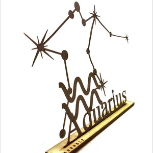 May include: A black metal cutout of the Aquarius constellation with the word "Aquarius" written below. The cutout is mounted on a small wooden base with the word "MADEHOLZ" printed on it.