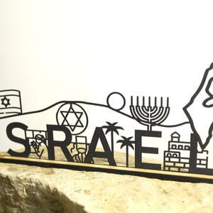 Puede incluir: Un letrero de madera negro cortado con láser que deletrea "ISRAEL" con símbolos icónicos y un contorno de mapa. El diseño incluye una bandera, la Estrella de David, una menorá, palmeras y un edificio. Montado sobre una base de madera clara.